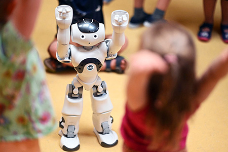 Ein kleiner weißer Roboter, der so gebaut ist, dass er menschliche Merkmale aufweist, hält die Arme über den Kopf. Im Vordergrund machen Kinder seine Haltung nach.