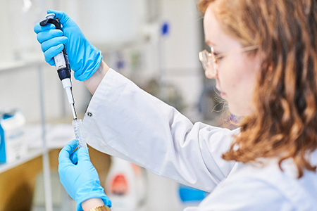 Eine Labortechnikerin mit Handschuhen und Laborkittel entnimmt mit einer Spritze eine Probe aus einem kleinen Behälter.