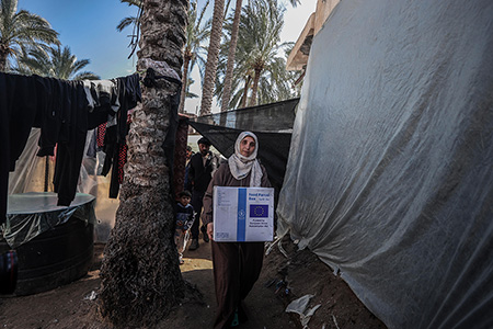 Eine Frau geht mit einem Paket zwischen behelfsmäßig mit Stoffbahnen konstruierten Zelten entlang. Auf dem Paket steht in Englisch „Nahrungsmittelpaket“ und „Finanziert mit der humanitären Hilfe der EU“. Zwei Männer und ein Kind folgen ihr. Im Hintergrund sieht man Palmen.