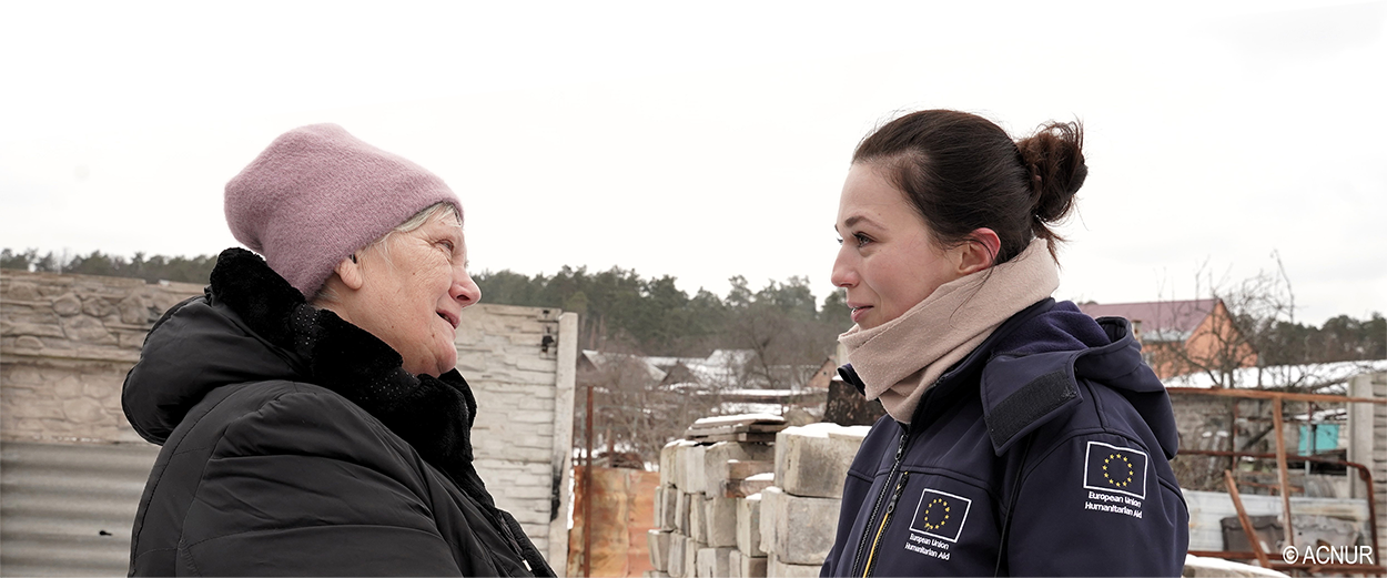 Lidiia e uma trabalhadora de ajuda humanitária da UE, identificável pela bandeira europeia na manga do casaco, conversam de mãos dadas numa zona com neve e gravemente atingida pela guerra.