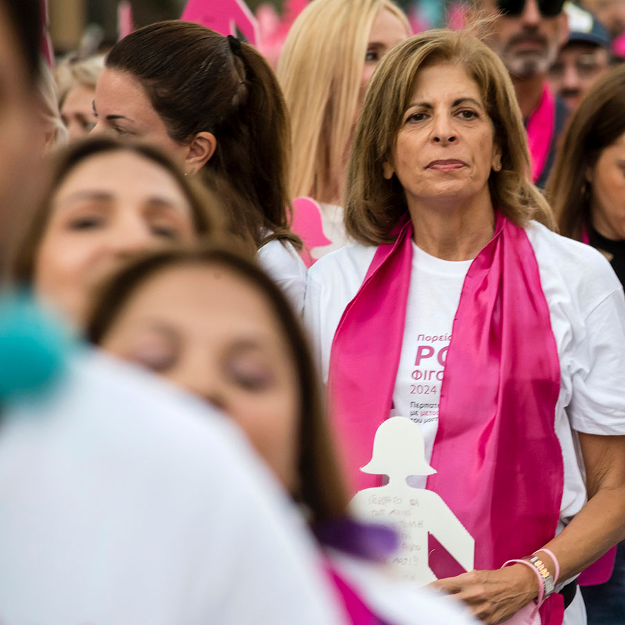 Stella Kyriakides går i et optog af mennesker iført lyserødt tørklæde og hvid T-shirt. Hun holder et udklip i form af en kvinde frem.