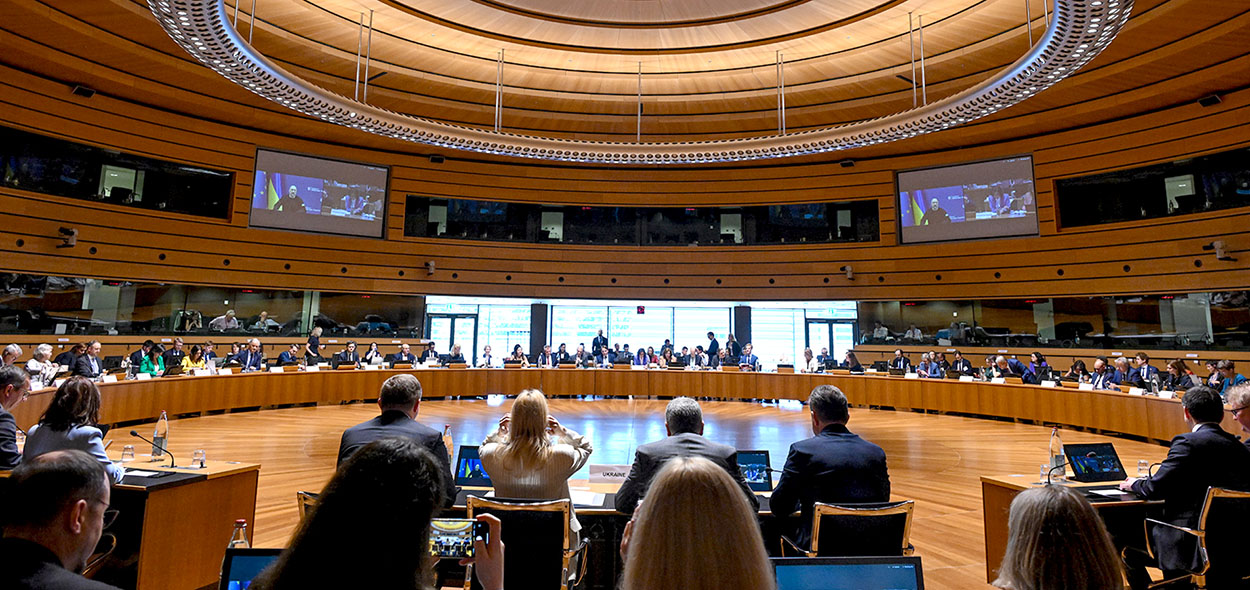 Varias personas están sentadas ante una mesa redonda, en una gran sala de reuniones. La sala está bien iluminada, es de arquitectura moderna y presenta grandes pantallas en las paredes.