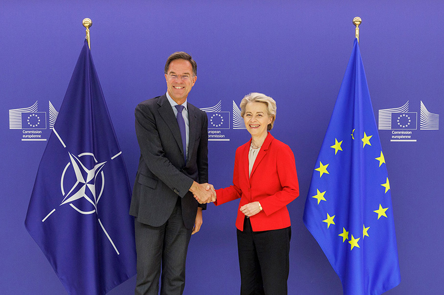 Ursula von der Leyen y Mark Rutte se estrechan la mano y sonríen, con una bandera de la OTAN a un lado y una bandera europea al otro. Al fondo, el logotipo de la Comisión Europea.