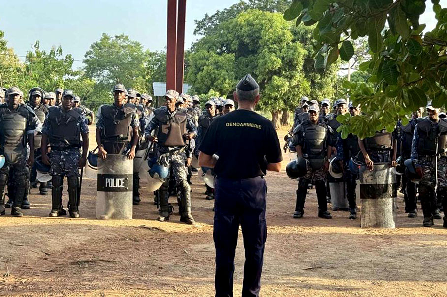 Un oficial de gendarmería, de espaldas a la cámara, habla con un grupo de agentes de policía ataviados con equipos de protección y escudos, que escuchan de pie en formación en el exterior, rodeados de árboles.