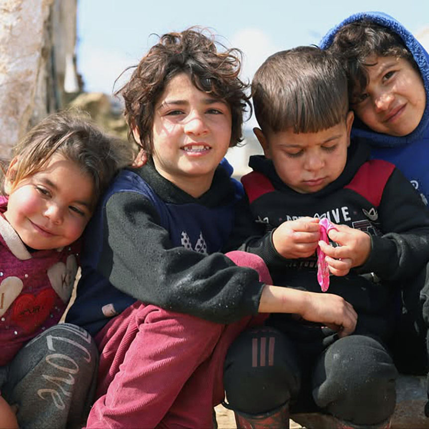 Cuatro niños sentados juntos al aire libre. Una se apoya en el niño de al lado y sonríe ligeramente, otro sonríe abiertamente a la cámara, el tercero mira con atención un objeto pequeño que tiene en las manos, y el cuarto se apoya en el tercero.