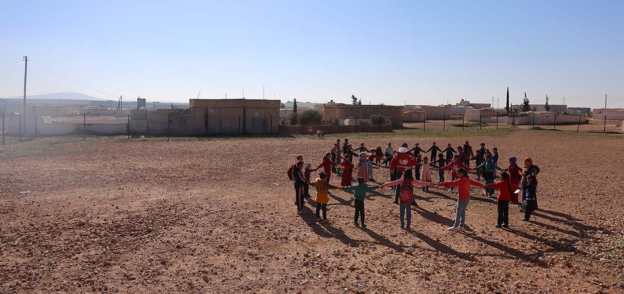 Varios niños unidos por las manos forman un gran círculo en un campo polvoriento. En el centro, un adulto vestido con una chaqueta roja y un emblema de media luna interactúa con ellos.
