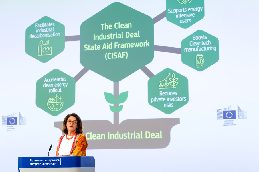 Teresa Riberová má prejav za rečníckym pultom. Za ňou je zobrazený diagram, v ktorého strede sú slová „the clean industrial deal State aid framework“ (rámec štátnej pomoci pre Dohodu o čistom priemysle). Stredná časť diagramu je spojená s piatimi bodmi: „facilitates industrial decarbonisation“ (uľahčuje priemyselnú dekarbonizáciu), „accelerates clean energy rollout“ (urýchľuje zavádzanie čistej energie), „reduces private investors risks“ (znižuje riziká pre súkromných investorov), „boosts clean tech manufacturing“ (posilňuje výrobu čistých technológií) a „supports energy intensive users“ (podporuje energeticky náročných používateľov). Pod diagramom sú slová „Clean Industrial Deal“ (Dohoda o čistom priemysle), ktoré označujú zdroj uvedených informácií, a zobrazené sú aj logá Európskej komisie.