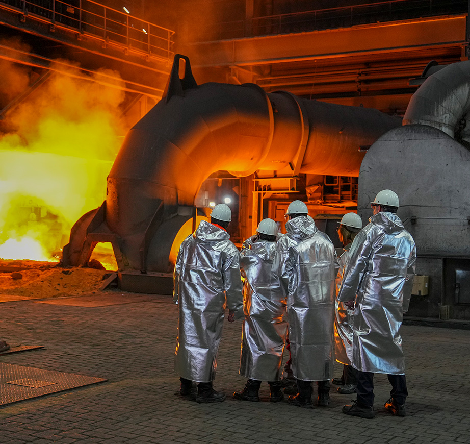 Skupina piatich ľudí v ochranných odevoch proti teplu si prezerá časť oceliarne.