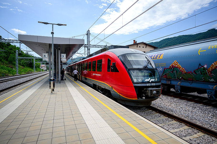 Na nástupišti železničnej stanice stojí červený vlak a vedľa neho je modrý nákladný vozeň.