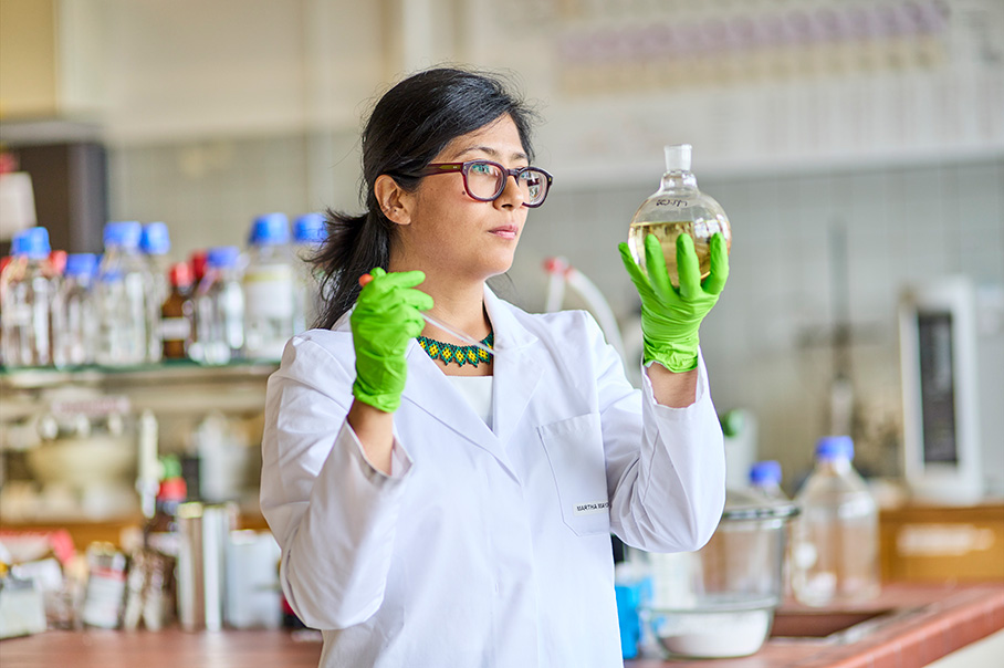 Dr. Martha C. Mayorquín-Torresová v bielom laboratórnom plášti drží v ruke banku s guľatým dnom a skúma jej kvapalný obsah, v druhej ruke má pipetu.