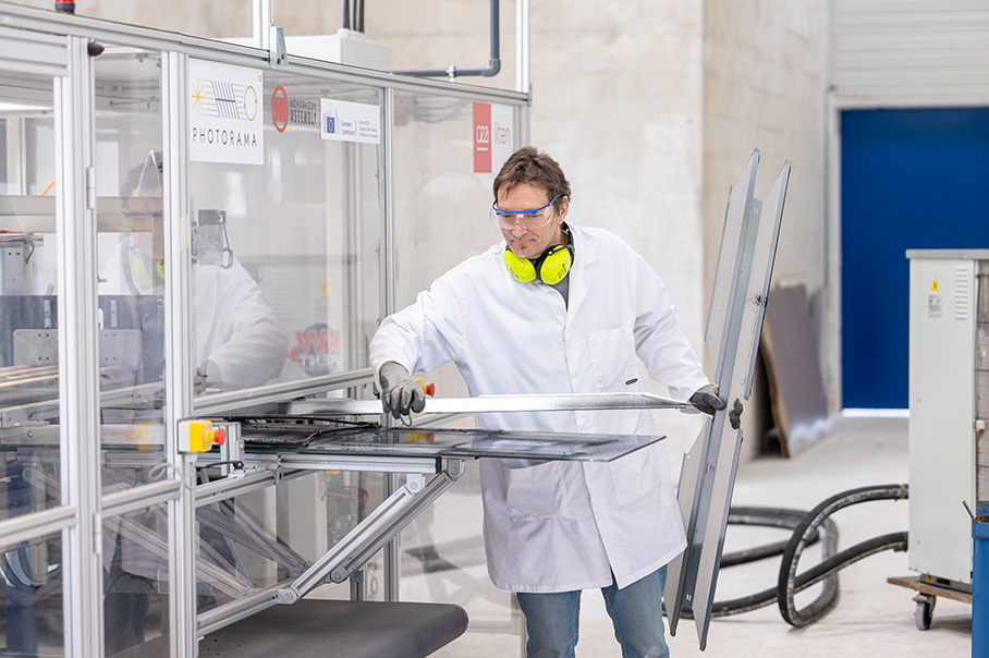 Muž v bielom laboratórnom plášti s chráničmi sluchu okolo krku vyberá zo stroja demontovaný starý solárny panel.