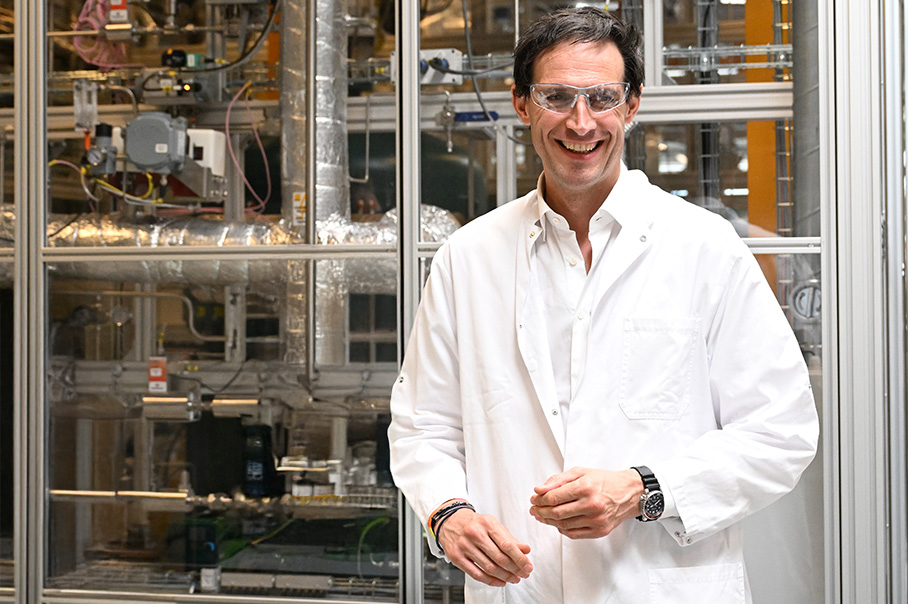 Wopke Hoekstra v bielom laboratórnom plášti stojí v priemyselnom zariadení, za ním sú kovové potrubia.