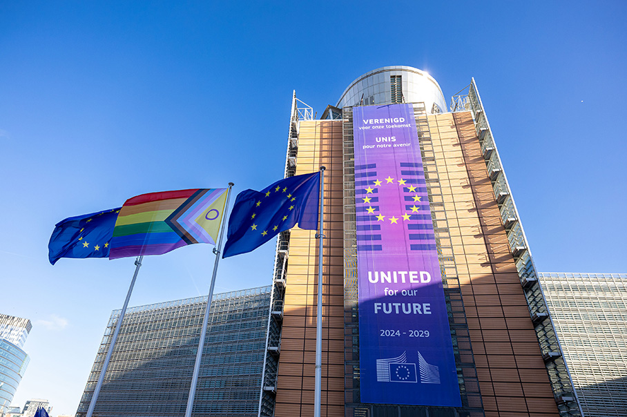 En Pride-flagga för intersexinkluderande utveckling och två EU-flaggor framför Berlaymont-byggnaden en klar och solig dag. På byggnaden hänger en banderoll med texten ”United for our future 2024–2029” (Förenade för vår framtid 2024–2029).