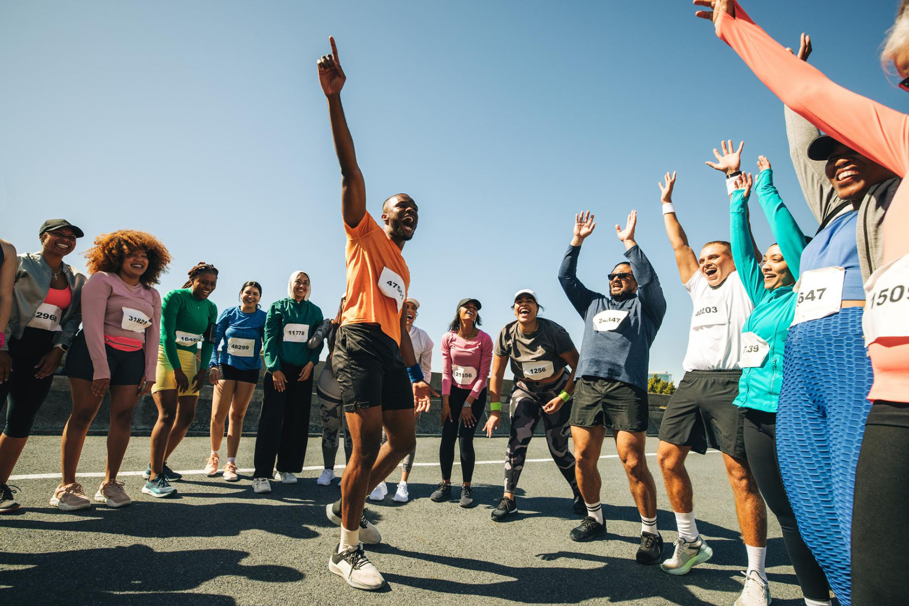 En grupp med olika män och kvinnor i löparkläder står i halvcirkel och jublar. En man står mitt i gruppen och har liknande löparkläder på sig, medan han triumferande pekar uppåt.