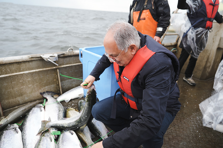 Costas Kadis hurkt neer op een boot op een grijze zee om de gevangen vis aan te raken. Hij draagt een reddingsvest.