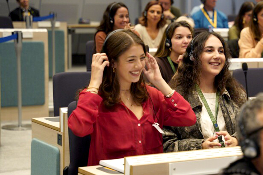 Ποικιλόμορφη ομάδα ατόμων που κάθονται μπροστά σε τραπέζια τοποθετημένα σε σειρά φορώντας ακουστικά. Σε πρώτο πλάνο, μια νεαρή γυναίκα γελά και προσαρμόζει τα ακουστικά στο κεφάλι της.