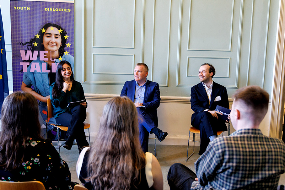 Ο κύριος Μάικλ ΜακΓκράθ κάθεται ανάμεσα σε δύο άλλα άτομα σε συζήτηση πάνελ, απευθυνόμενος σε ένα νεανικό ακροατήριο. Μια αφίσα στο πίσω μέρος αναγράφει τις λέξεις «We talk» (Ας μιλήσουμε).