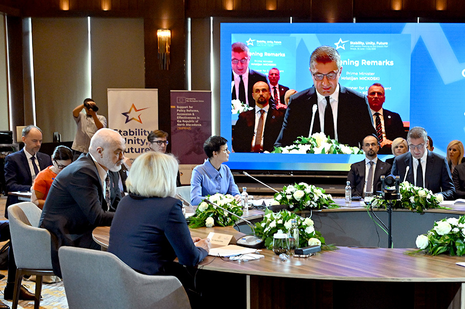 Марта Кос и други седят на кръгла конферентна маса. Образът на оратора се прожектира върху екран на заден план. Други седят в периферията на основната маса.