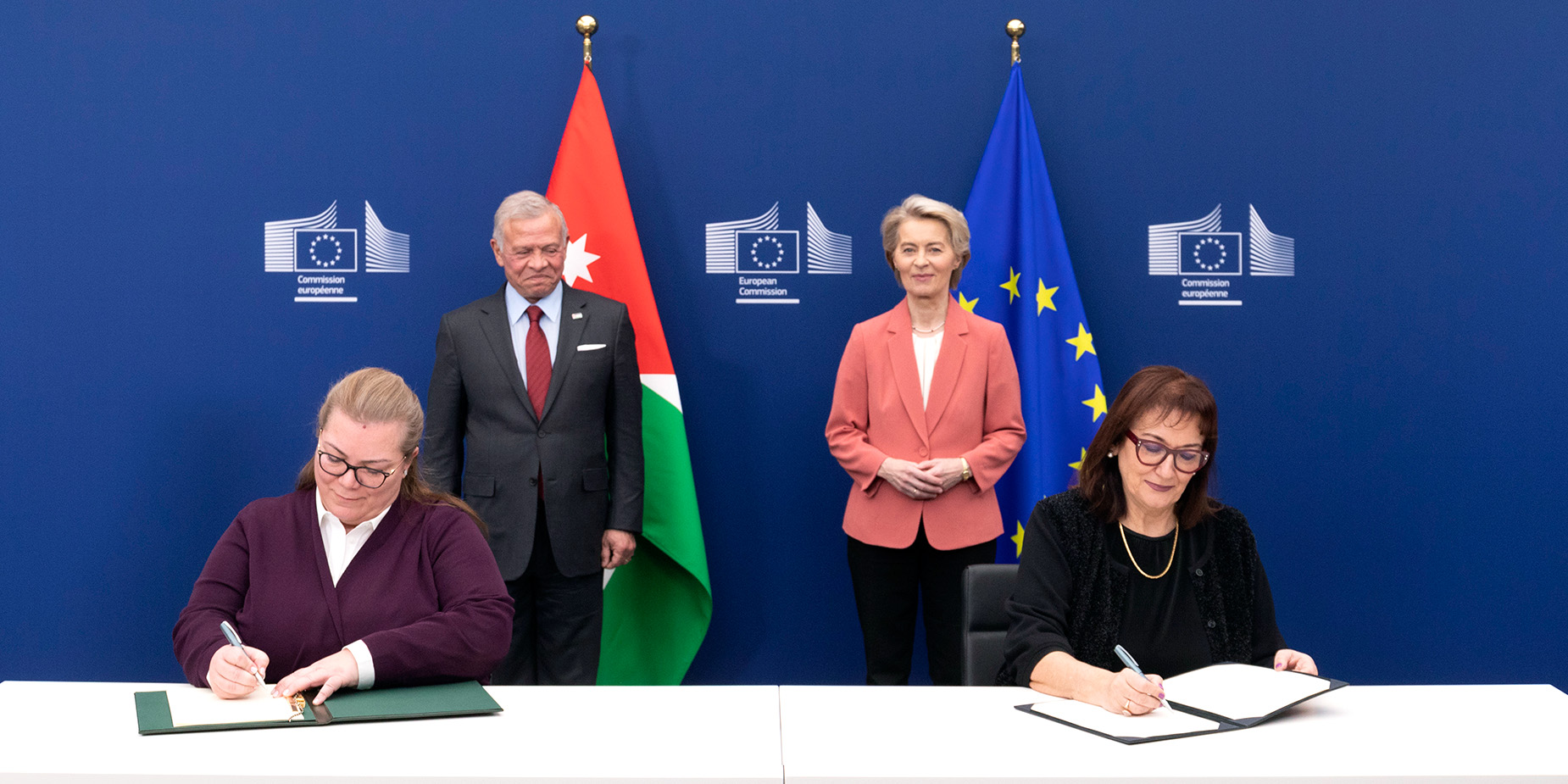 Дубравка Шуица и Зейна Тукан седят на маса, всяка от тях подписва документ. Зад тях са застанали Урсула фон дер Лайен и Абдула II, крал на Йордания. Зад тях са разположени йорданско и европейско знаме. Синият фон включва три логотипа на Европейската комисия.