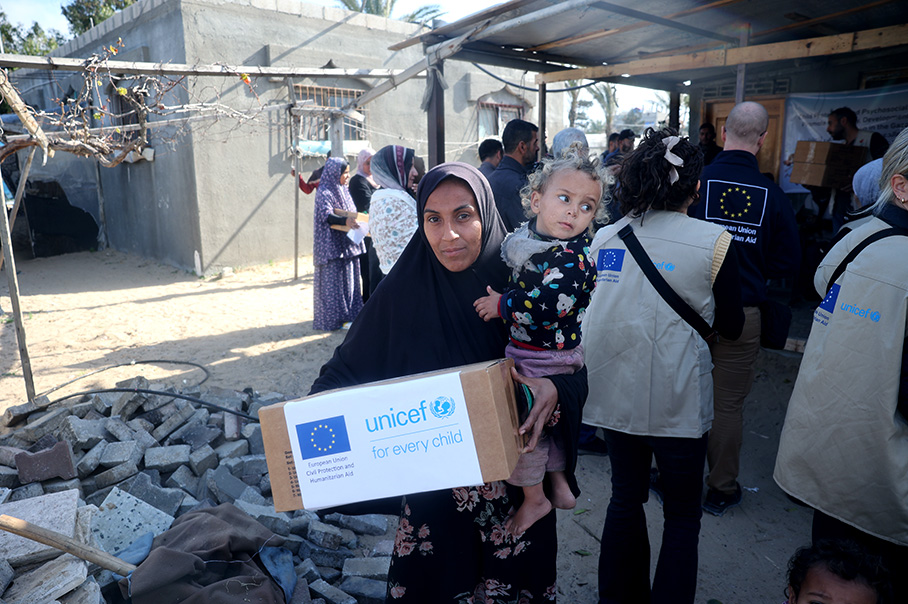 Жена от Газа с дете на ръце държи кутия, на която е изобразено европейско знаме и са изписани думите „УНИЦЕФ за всяко дете“. Зад нея стои група хора, някои от които носят жилетки със същите символи. По улицата има отпадъци.