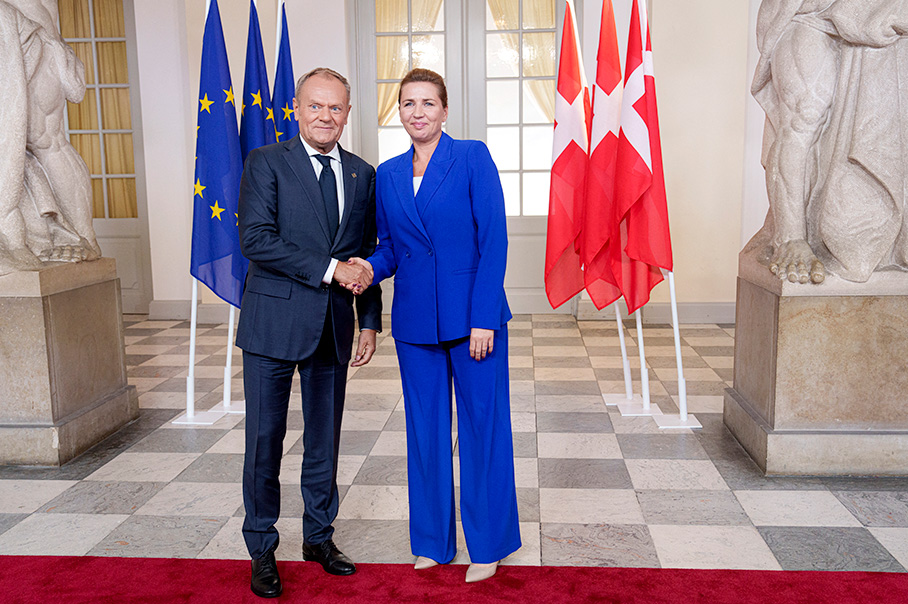 Доналд Туск и Мете Фредериксен са застанали прави и се ръкуват в помещение с бели стени. На заден план се виждат мраморни статуи от двете страни. Зад тях са поставени множество знамена на Европейския съюз и Дания.