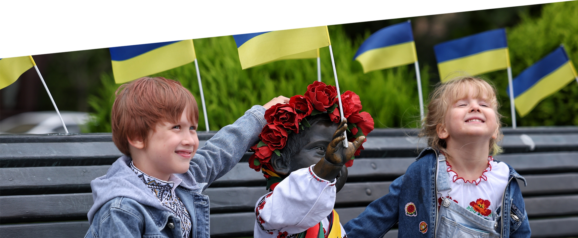 Deux enfants assis sur un banc de part et d’autre d’une statue vêtue de la chemise traditionnelle ukrainienne vyshyvanka, souriant et posant pour des photographies.