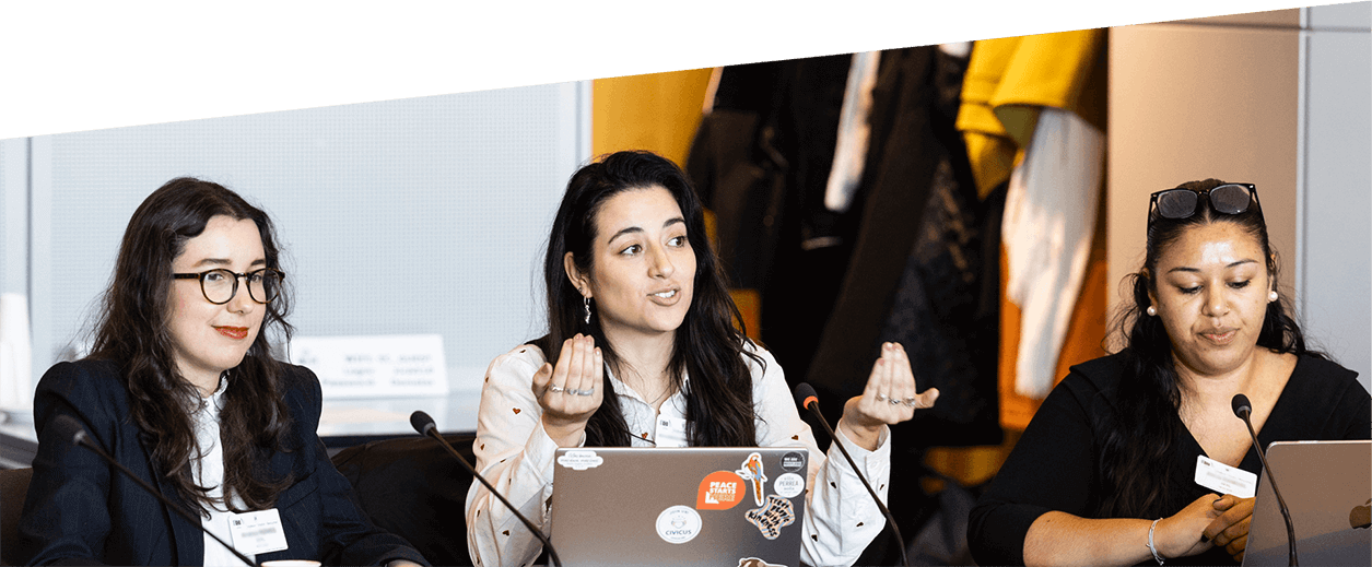 Tre unga kvinnor sitter vid ett konferensbord. Alla tre har en mikrofon framför sig och två har sina bärbara datorer öppna.