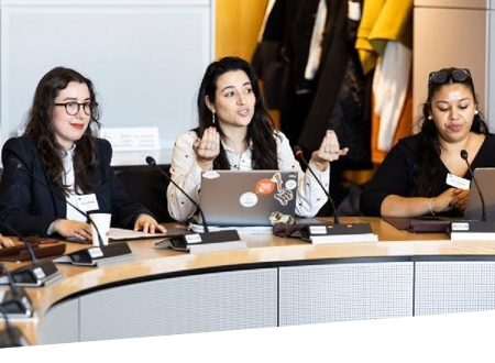 Trei tinere sunt așezate în spatele unei mese de conferință curbate. Fiecare are un microfon în față, iar două dintre ele au laptopuri deschise.