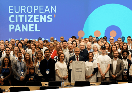 O sală de conferințe plină de participanți, în picioare în fața unui ecran care afișează cuvintele „European Citizens’ Panel” (Grupul de dezbatere al cetățenilor europeni).