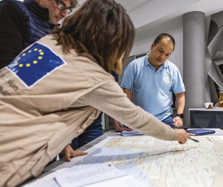 Vier Mitarbeiter in einer humanitären Koordinierungsstelle schauen gemeinsam auf eine Landkarte der Ukraine. Eine von ihnen trägt eine Jacke mit der EU-Flagge auf dem Rücken und zeigt auf die Karte.