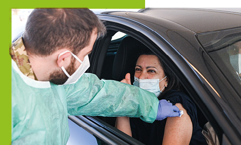 Медицинский работник в защитном снаряжении вводит женщине вакцину через окно автомобиля.