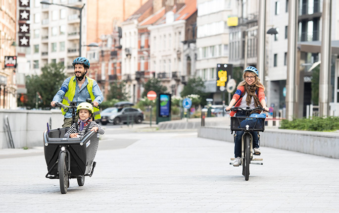 Uma mulher passeia de bicicleta por uma cidade ao lado de um homem numa bicicleta de carga com uma criança no assento da frente, os três usam capacetes.