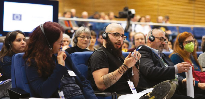Eine Gruppe von Bürgerinnen und Bürgern sitzt mit Kopfhörern in einem Auditorium, während ein junger Mann in der vorderen Reihe spricht.
