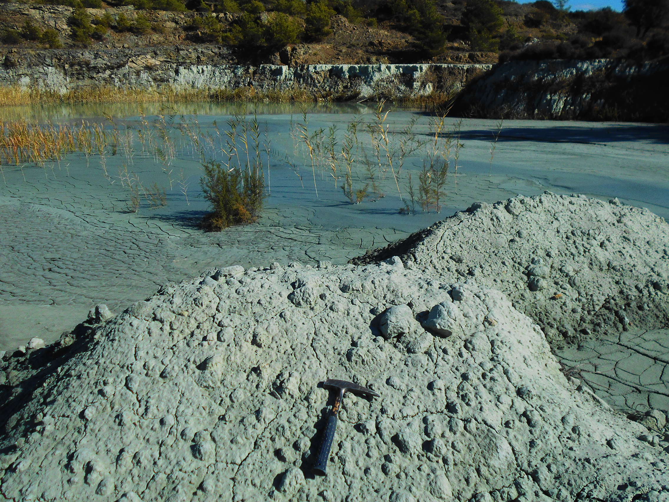 A geological hammer rests on a mound of pale gray sediment beside a shallow pond with reeds and cracked mud, surrounded by rocky terrain.